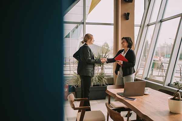 Two professionals shaking hands while holding documents in a modern office with large windows.