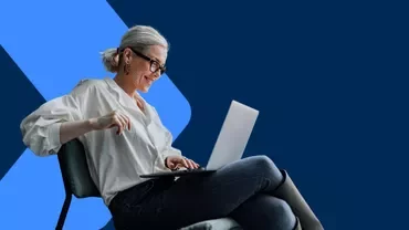 Person working on a laptop while seated on a chair against a blue background.