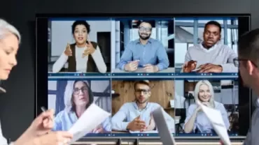 Professionals participating in a hybrid meeting with colleagues joining via video conference on a large screen.
